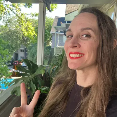 Amanda Bouldin smiling on a sunlit porch with a peace sign, surrounded by green plants and trees